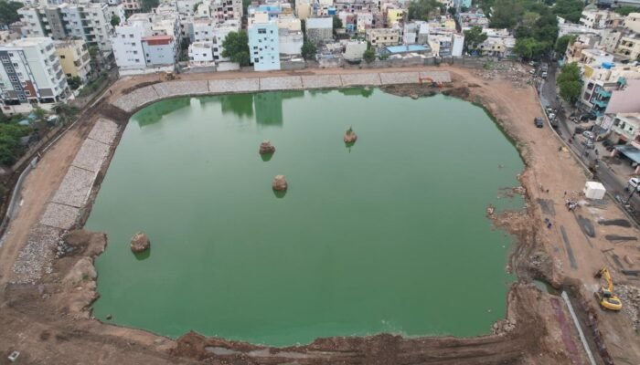 Aerial view of a large, square pond, identified as a Bathukamma kunta, with construction visible around the edges in an urban area.