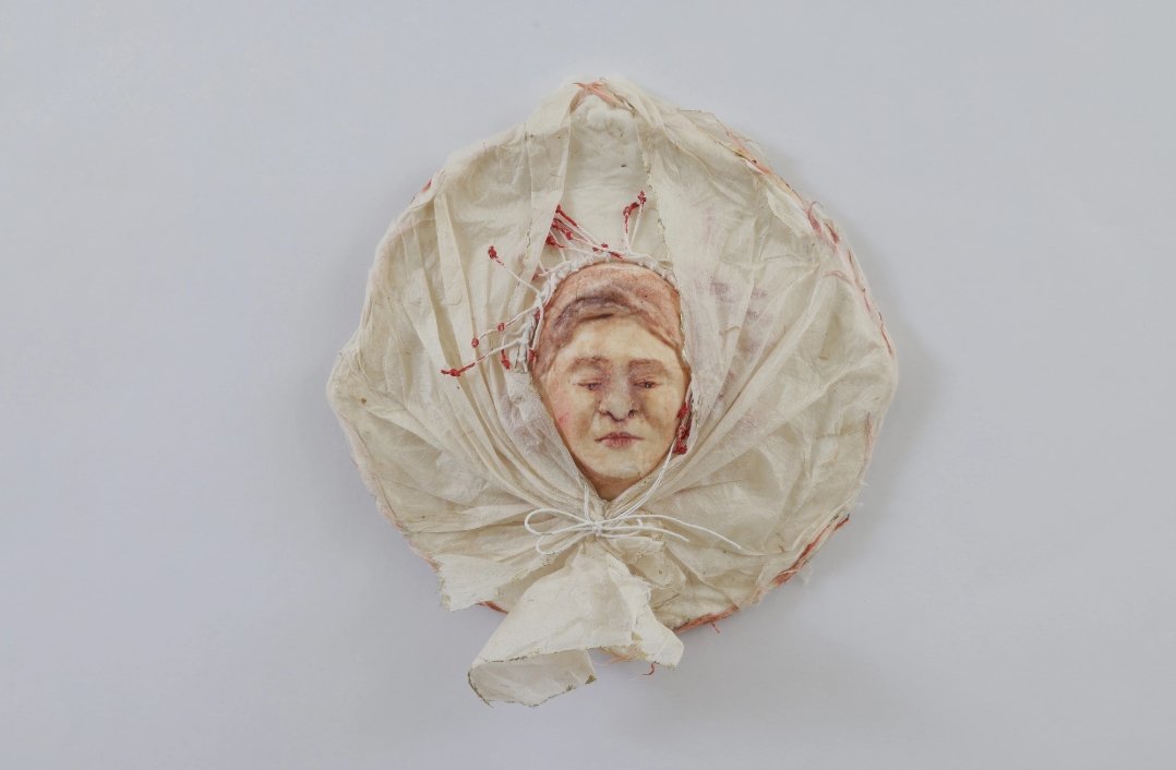A circular bundle of fabric with red threads surrounds and shelters a figure, illustrating the concept of “a room for refuge” through artful protection